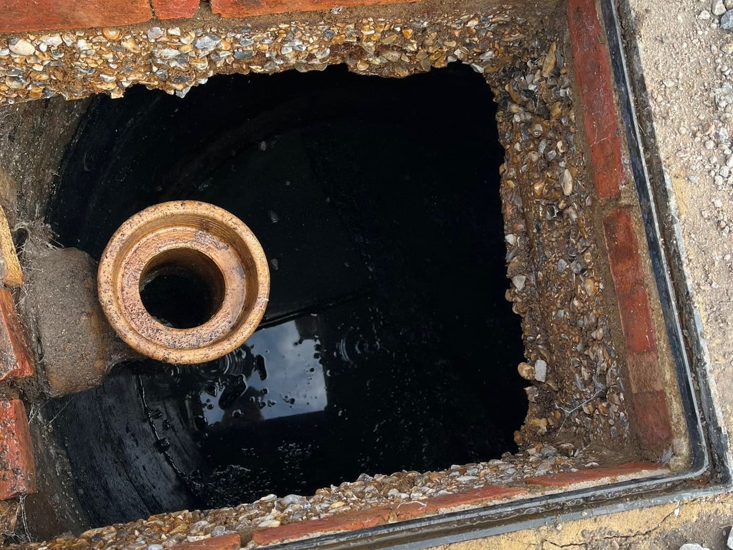 Looking down into an open brick inspection chamber with a clay pipe socket and dark water below, a gloved hand at the edge