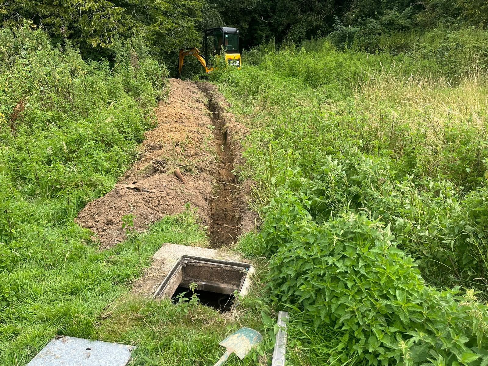 Trench dug through rough ground to an open chamber, with the excavator still on site