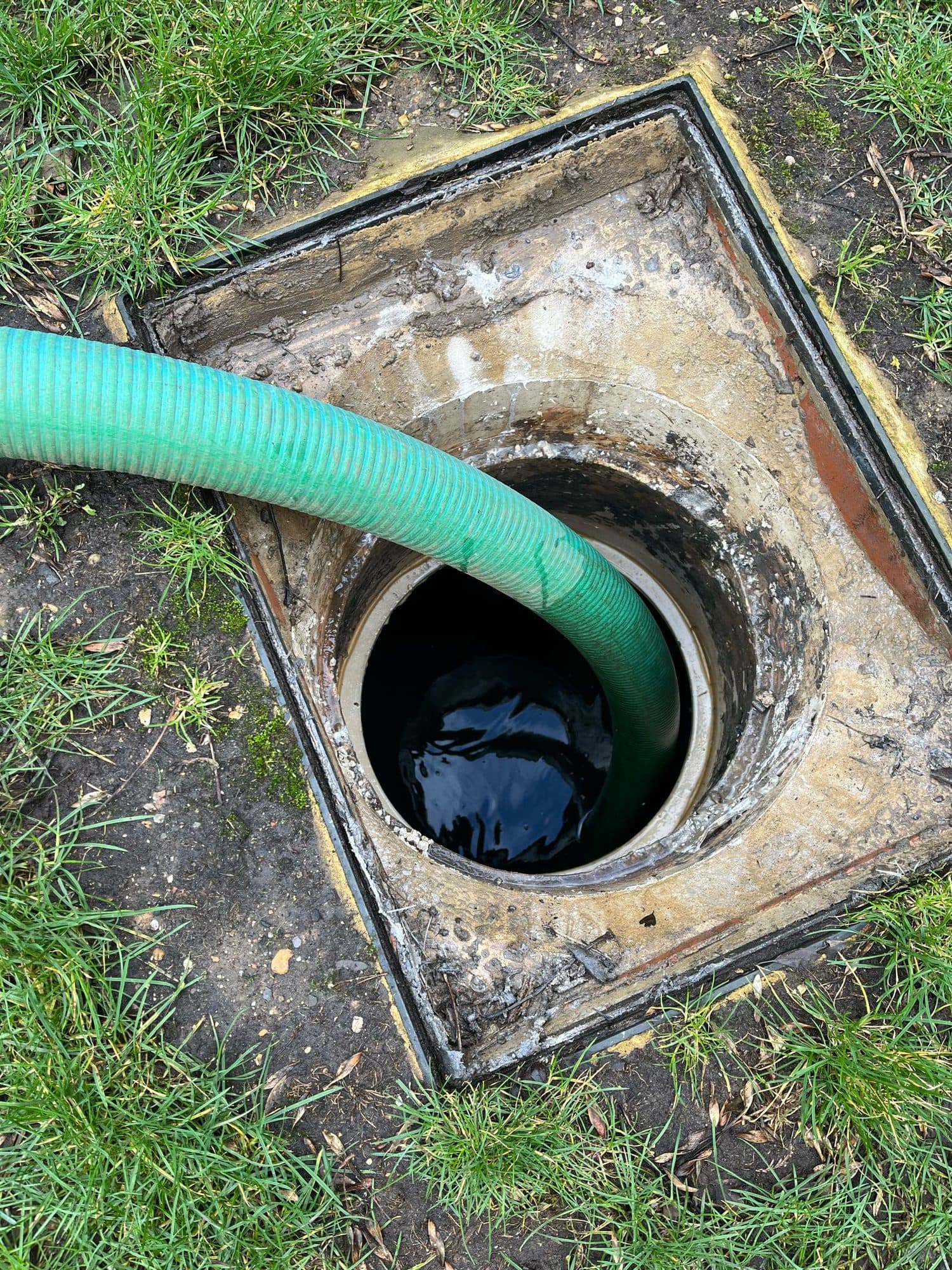 Green suction hose lowered into a square inspection chamber set in a lawn